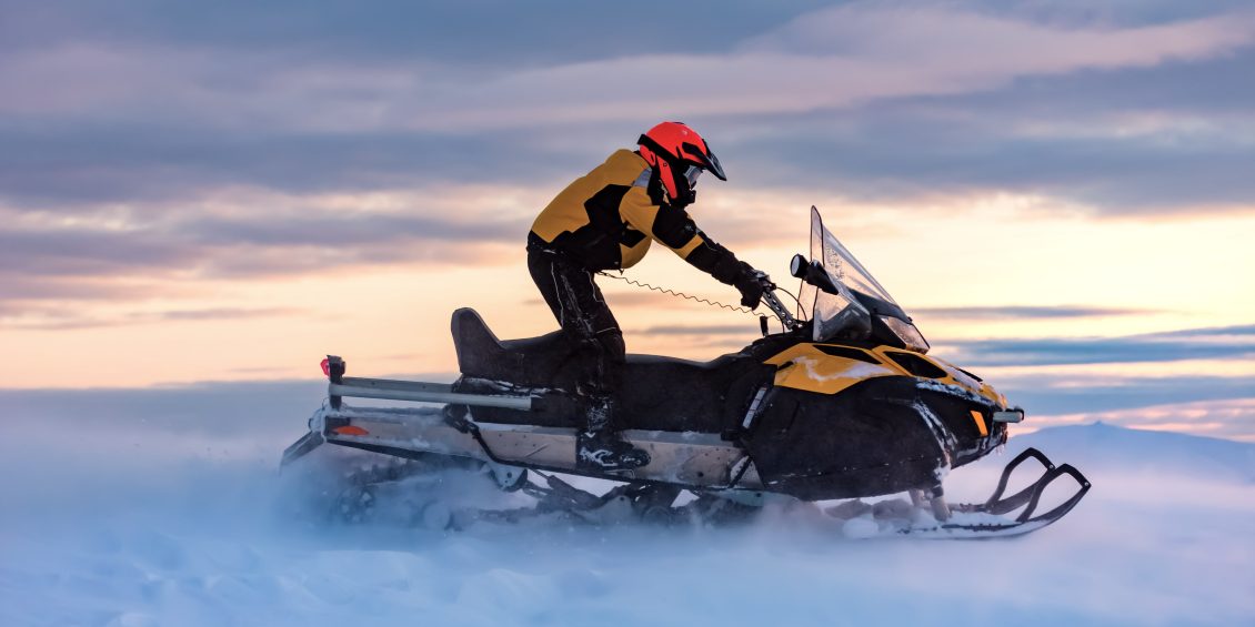 Rider in a yellow suit speeding through the snow on a snowmobile at sunset.